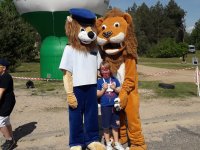 Maskotki policyjne, po lewej stronie niebieski, a po prawej stronie brązowy lew. Pomiędzy maskotkami stoi dziecko- uczestnik zabaw.