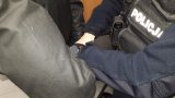 Umundurowany policjant podczas doprowadzenia zatrzymanego, zdejmuje mu kajdanki założone na ręce trzymane z tyłu.