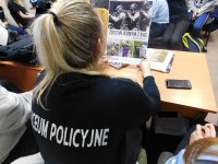 Uczennica ubrana w czarną koszulkę z napisem na plecach ,,LICEUM POLICYJNE&quot;. W ręku trzyma plakat promujący służbę w Policji.