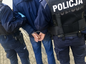 Policjanci podczas doprowadzenia osoby zatrzymanej, która ma założone kajdanki na ręce trzymane z tyłu.